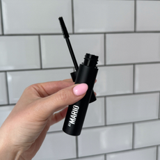 Close up of a handheld view of the Makeup by Mario Master Mascara open with its wand in front of a white tiled background