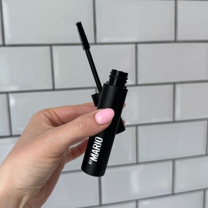 Close up of a handheld view of the Makeup by Mario Master Mascara open with its wand in front of a white tiled background