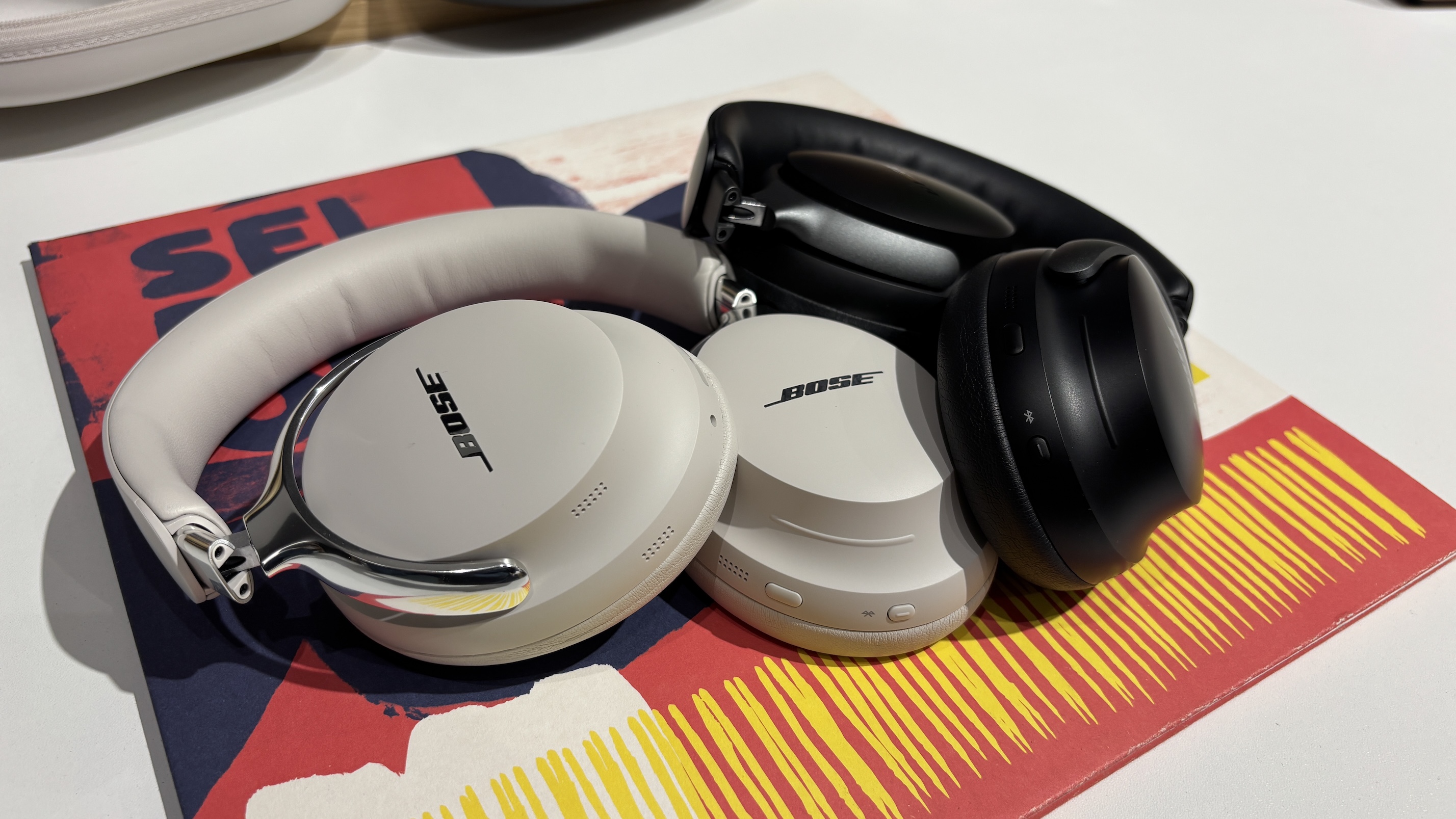 A pair of Bose QuietComfort Ultra Headphones (2nd Gen) and their first-gen predecessors lying on a record sleeve on a white surface.