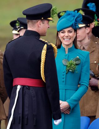 Kate Middleton wearing a teal coat and hat laughing as Prince William, wearing a military uniform, stands with his back to the camera in front of her on St. Patrick's Day 2023