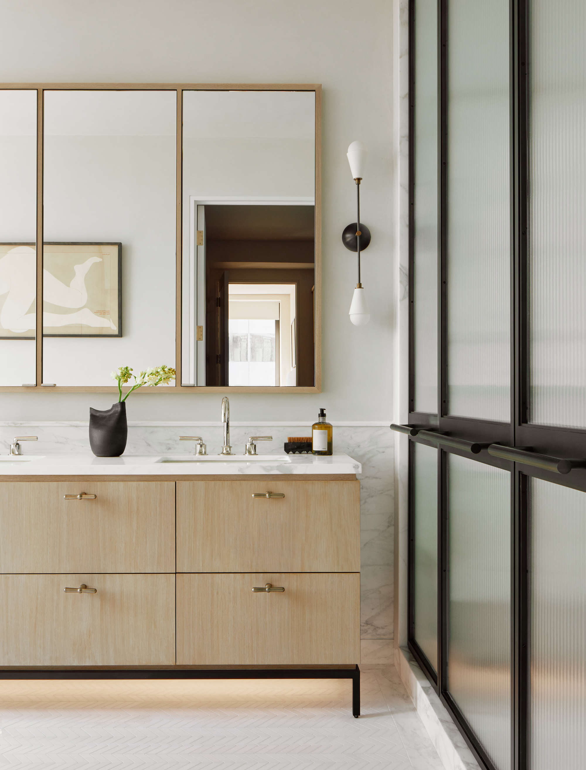 neutral bathroom with white floors, fluted glass doors with black accents, timber vanity with white marble counter, double sinks, silver taps, vase of flowers, mirrored medicine cabinets, wall sconce, and white walls
