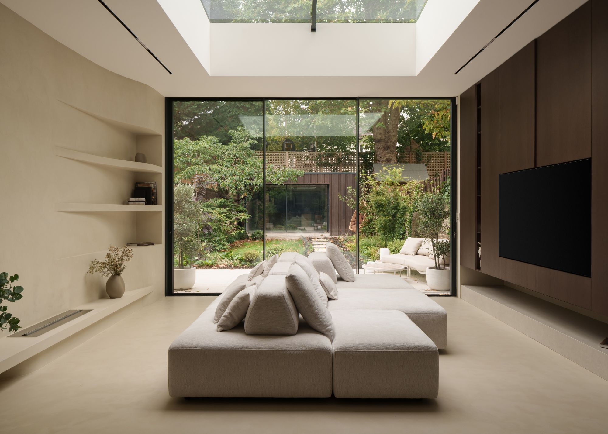 A modern open plan living space with a large skylight above a central square cream sofa in front of sliding glass doors with seating on both sides; open built-in shelving on one, and a tv on the other