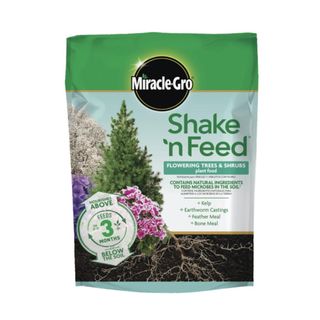 A bag of Miracle-Gro shake 'n feed with a dark green stripe at the top and a picture of trees and soil with a clear blue sky below it