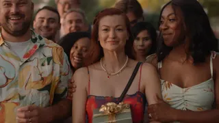 A woman (Leah McKendrick) holds a gift while standing at the front of a crowd, with another woman (Ego Nwodim) holding her arm and smiling at her, in 'Scrambled.'