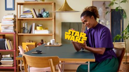 A smiling woman holds up the Lego Brick-Built Star Wars Logo set whilst sat at a table