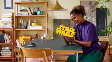 A smiling woman holds up the Lego Brick-Built Star Wars Logo set whilst sat at a table