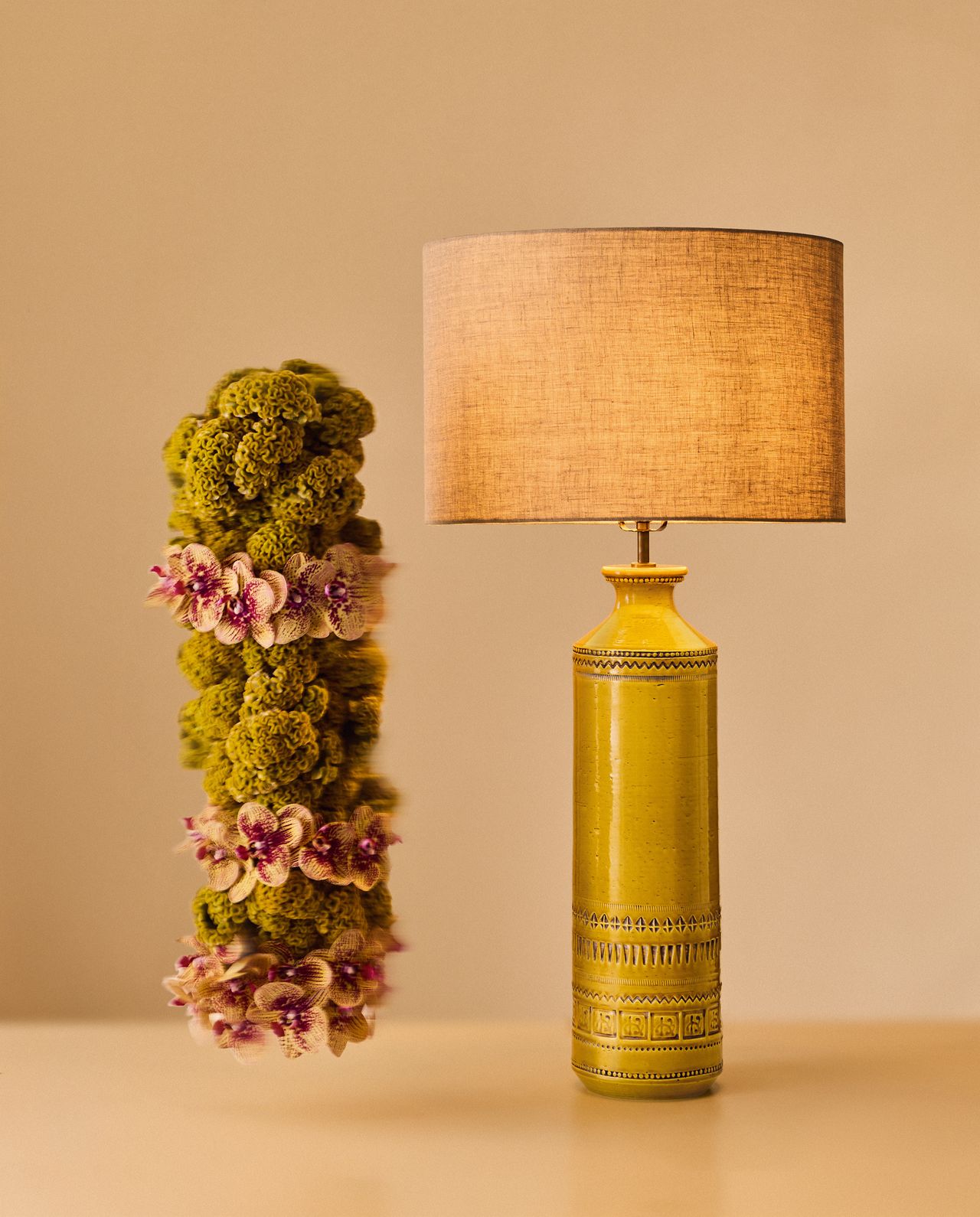 Image of an artisanal ceramic, yellow lamp next to an artisanal foliage structure in a display setup that is drenched in a beige color