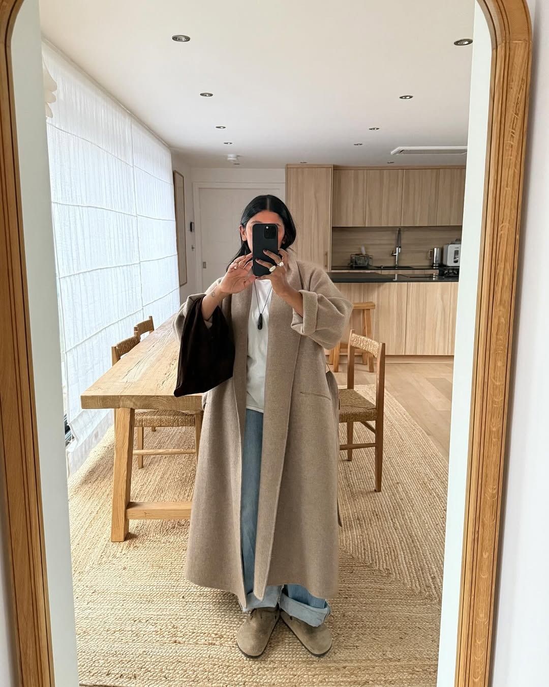 Influencer Monikh wears a long beige coat with cuffed light-wash jeans, a white crewneck, and Birkenstock Boston clogs.