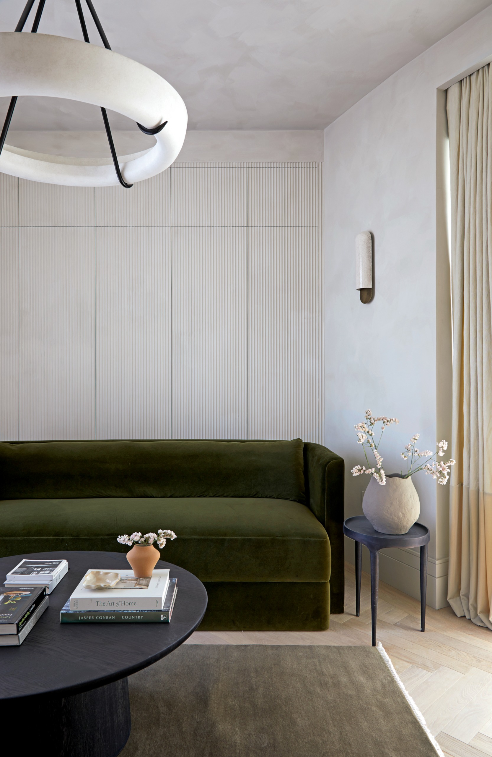 Snug with plaster-texture white walls, fluted storage, olive green velvet sofa, olive rug, round coffee table and white ring pendant light