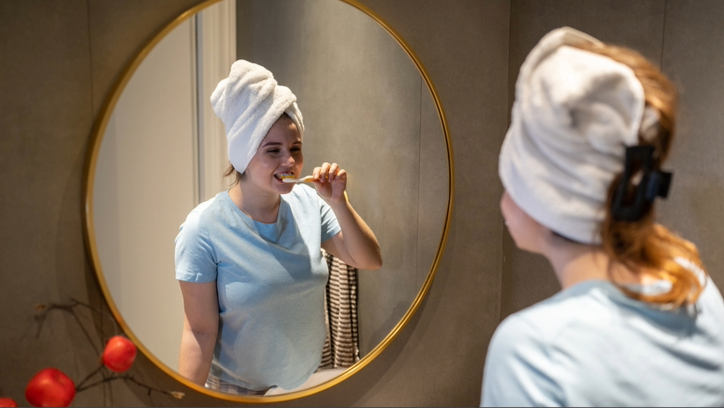 a woman with white towel on head brushing teeth in mirror after shower before bed