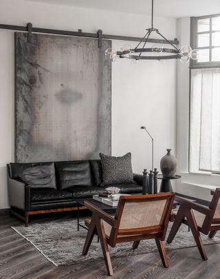 A sitting area with a black leather couch, two wooden chairs, a coffee table, a rug and a wall painting.