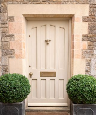 cream panelled front door on older stone property with gold letterbox and bee shaped door knocker, plus green round box plants on either side of door