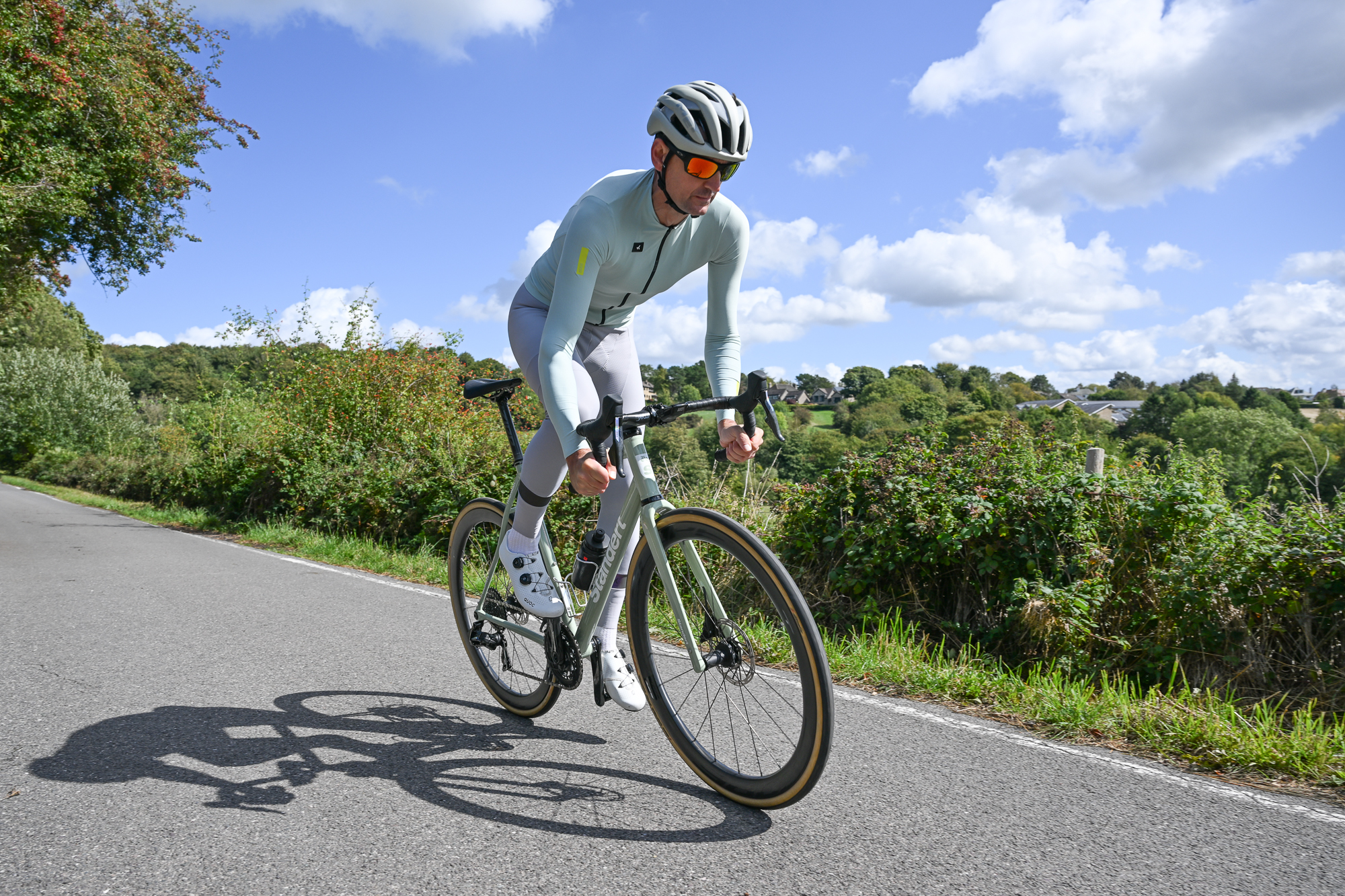 Man riding out of the saddle from left to right wearing pale green long sleeved jersey and pale grey bib tights