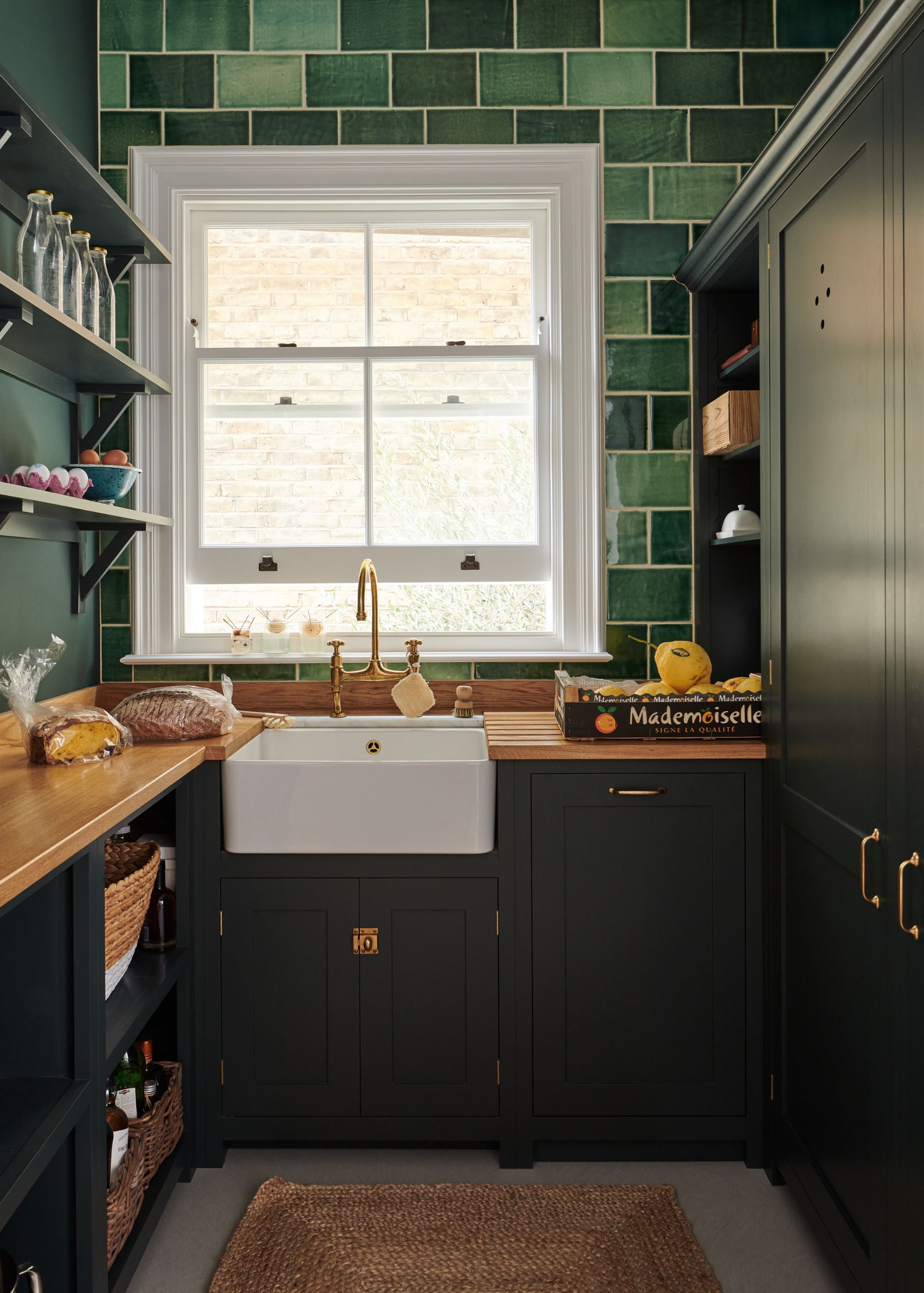 A shaker style kitchen with deep green cabinets and a mixed colored green backsplash leading towards the ceiling. There is also a large window slightly perched open beside a sink