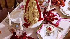 close up of table set for christmas with white plates, silver candlesticks and white candles, a patterned tablecloth, stacked dessert, red ribbons and grapes