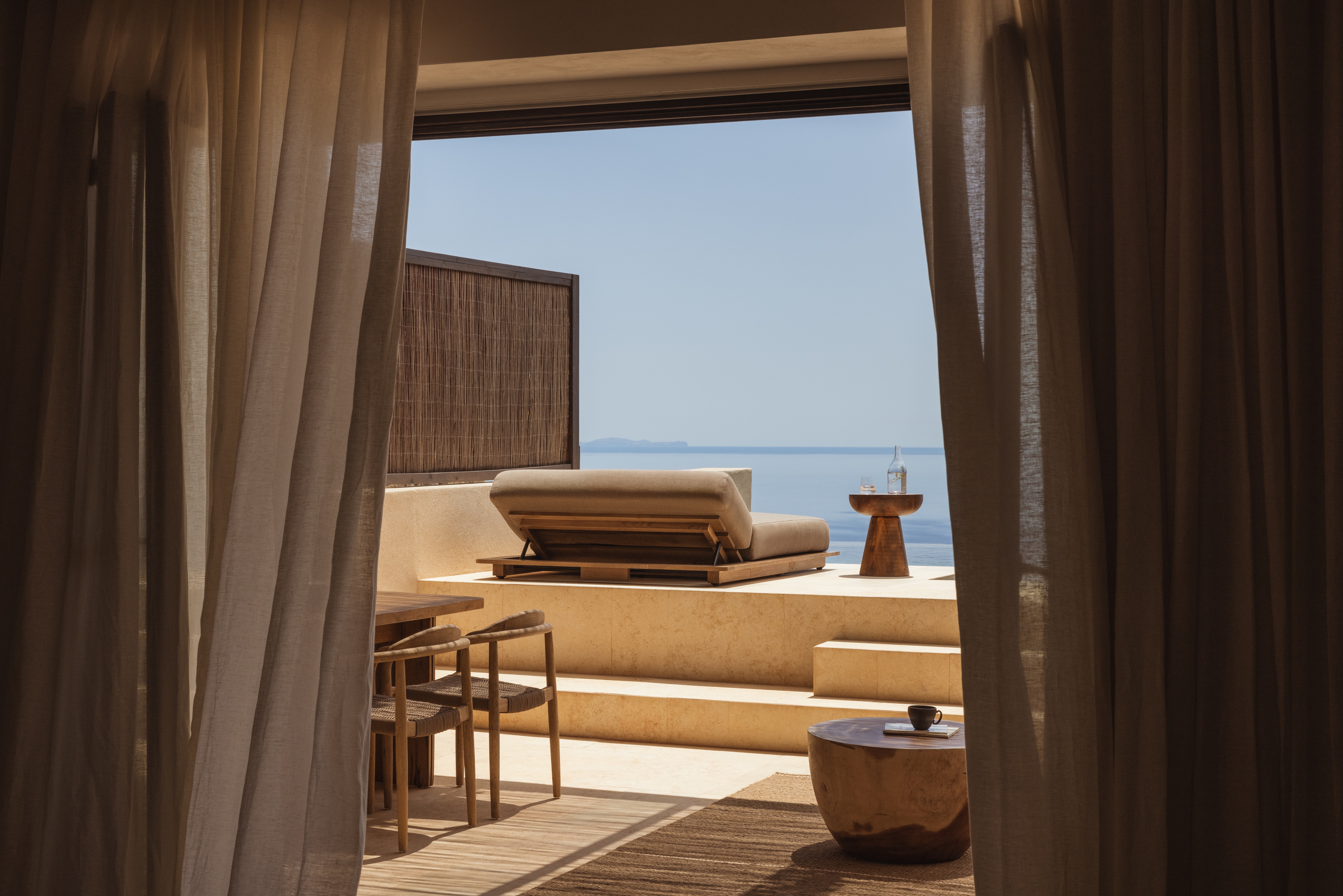 A hotel room in tones of beige. Gauzy curtains lead out onto a stone terrace with chairs, a side table and day bed