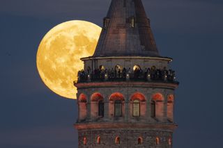 full moon behind a large tower with people standing on a viewing platform at the top