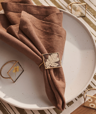 square gold-rimmed napkin rings with different pressed flowers inside on a brown linen napkin