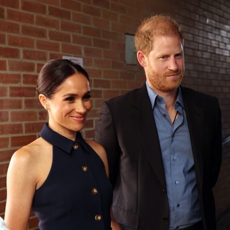 Meghan Markle wears a navy dress and Prince Harry wears a blue shirt with a dark suit, and Lord Ivar Mountbatten smiles while wearing a gray suit