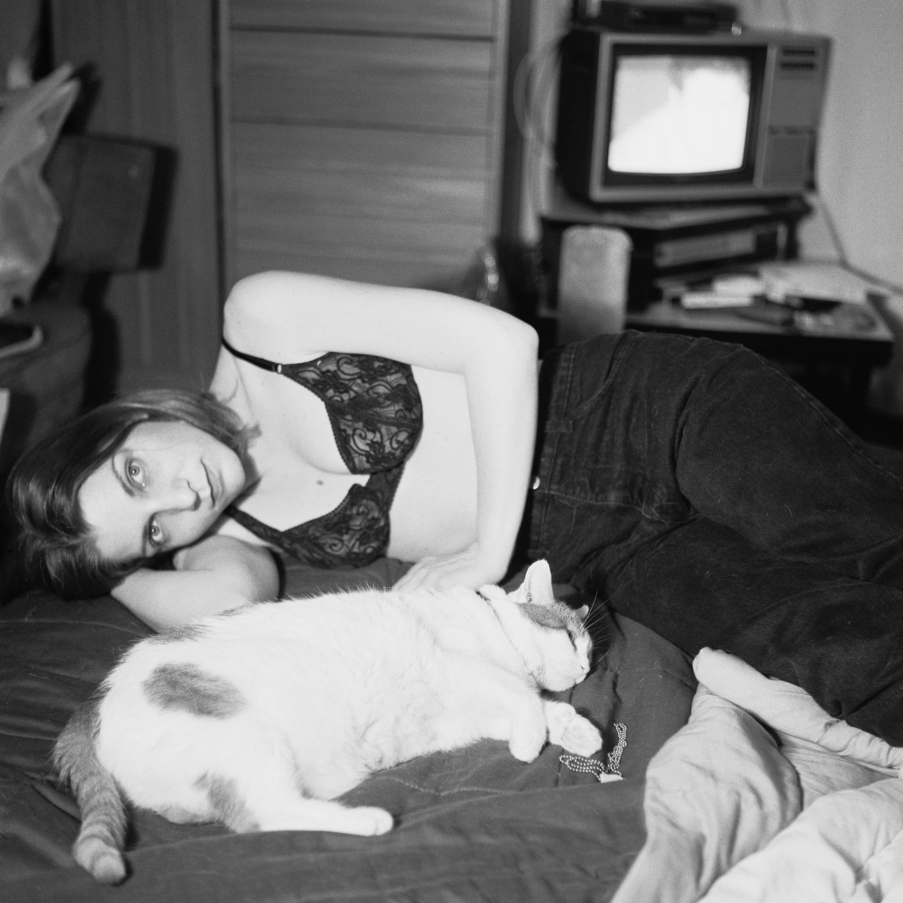 A person lounges on a bed in casual attire, with a white cat sleeping beside them, in a cozy indoor setting. A television is visible in the background