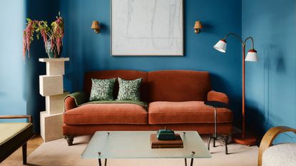A living room with blue color-drenched walls, a rust-colored sofa and modern decor, and a beige carpet.