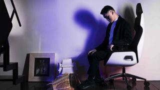 A man sits on a white chair with an ominous shadow behind him, staring blankly at some architectural models.