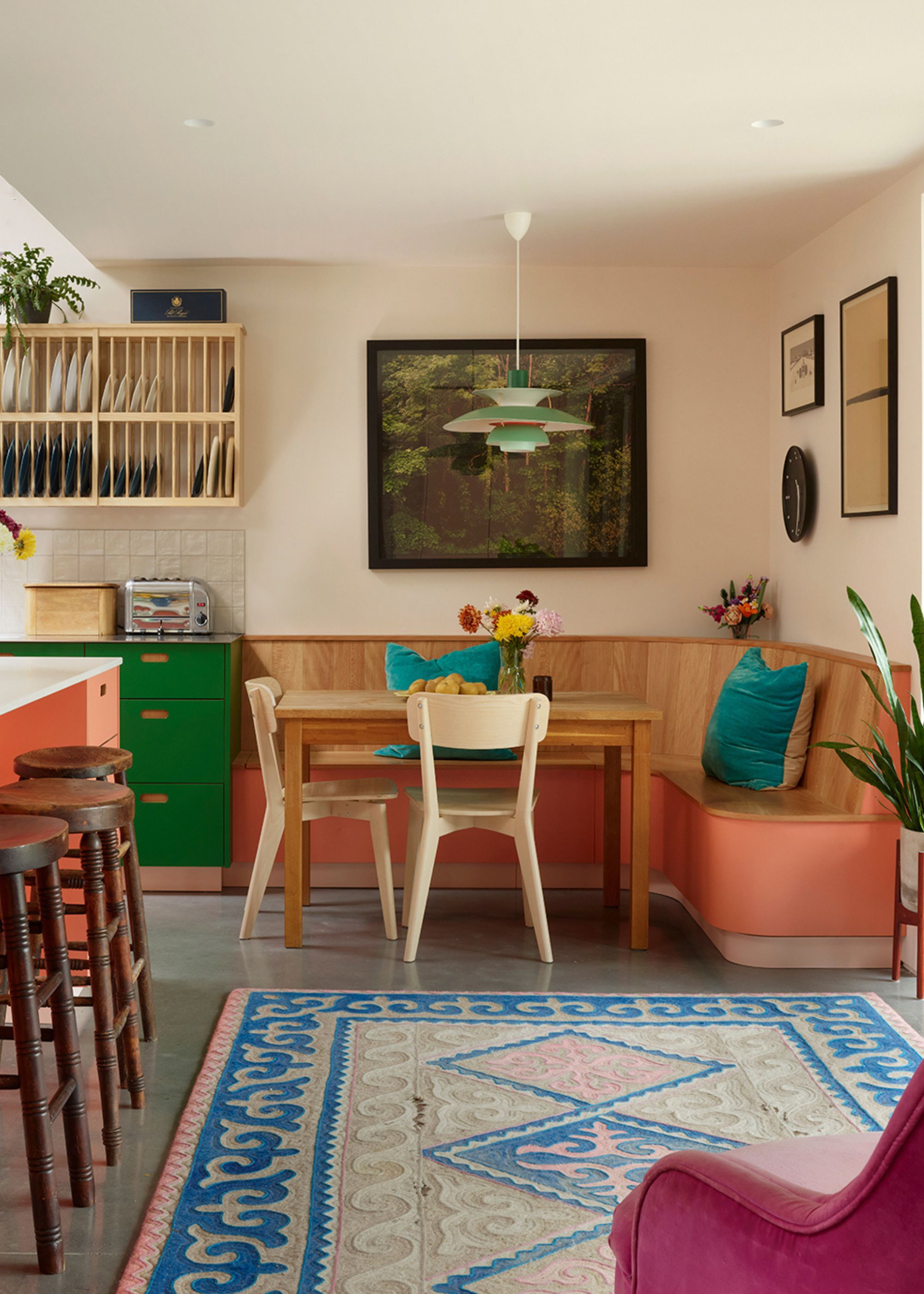 A colorful kitchen with banquette seating with framed pictures and art work hanging above it. Next to the dining area is also a rack of plates as well as green colored cabinets and drawers.