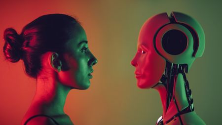 A woman with a pony tail looks to the right at a robot face looking back at her with red and green lighting in the background.