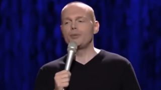 Bill Burr telling a joke on stage in a black shirt