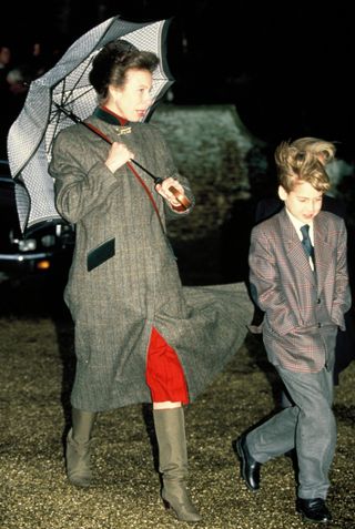 Princess Anne wearing a long gray coat and holding an umbrella walking next to a young Prince William in a. suit and tie on Christmas