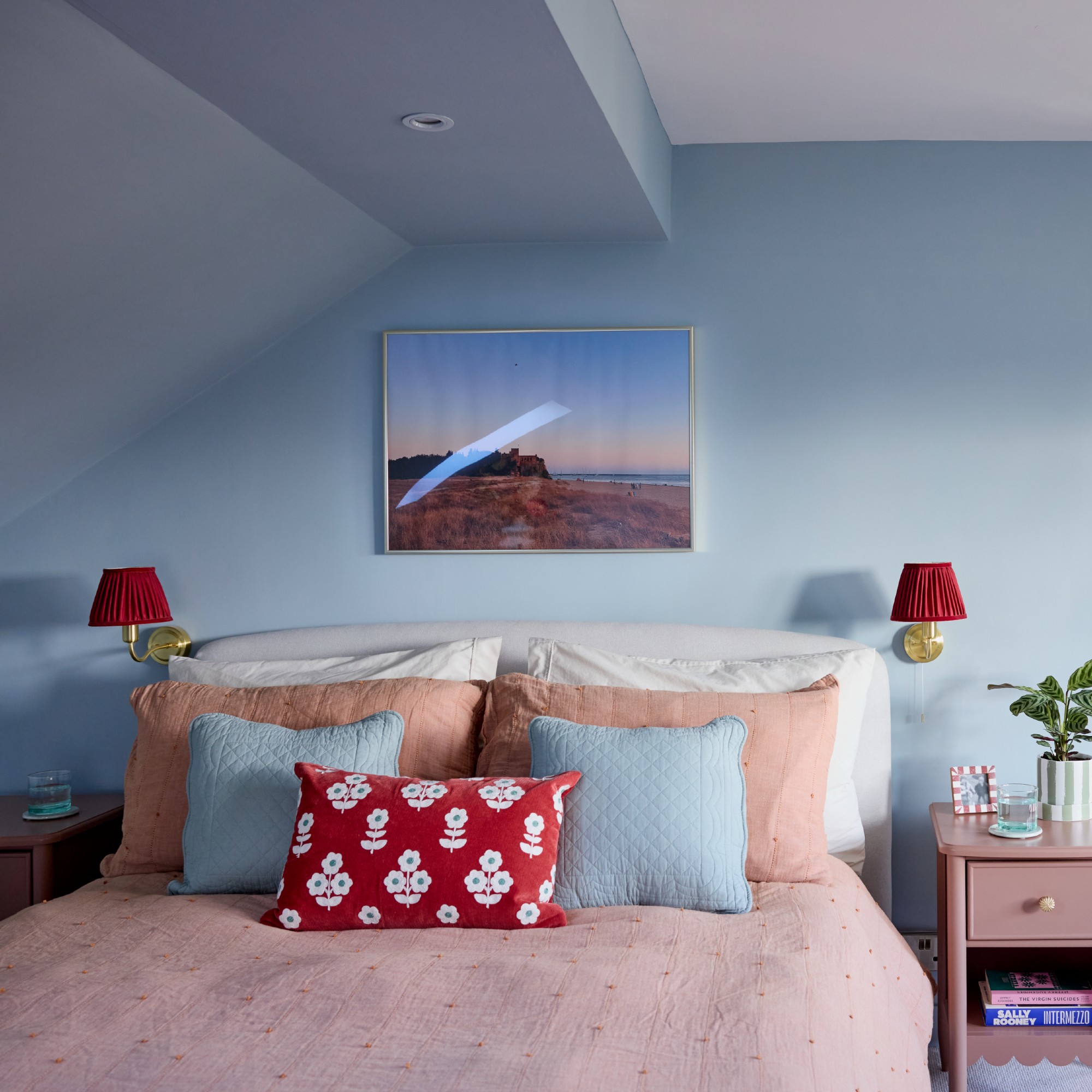 A soft blue-painted bedroom with a slanted ceiling, pink bedding and bedside tables with brass wall lights with red lampshades above them