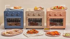 the new new Our Place Wonder Ovens photographed side by side on a taupe background showing the blue, cream and pink new oven designs surrounding by plates of food