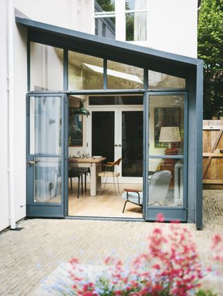 stylish grey and glass orangery added onto the side of a house, leading onto a tiled garden area, with a wooden dining set and chair inside