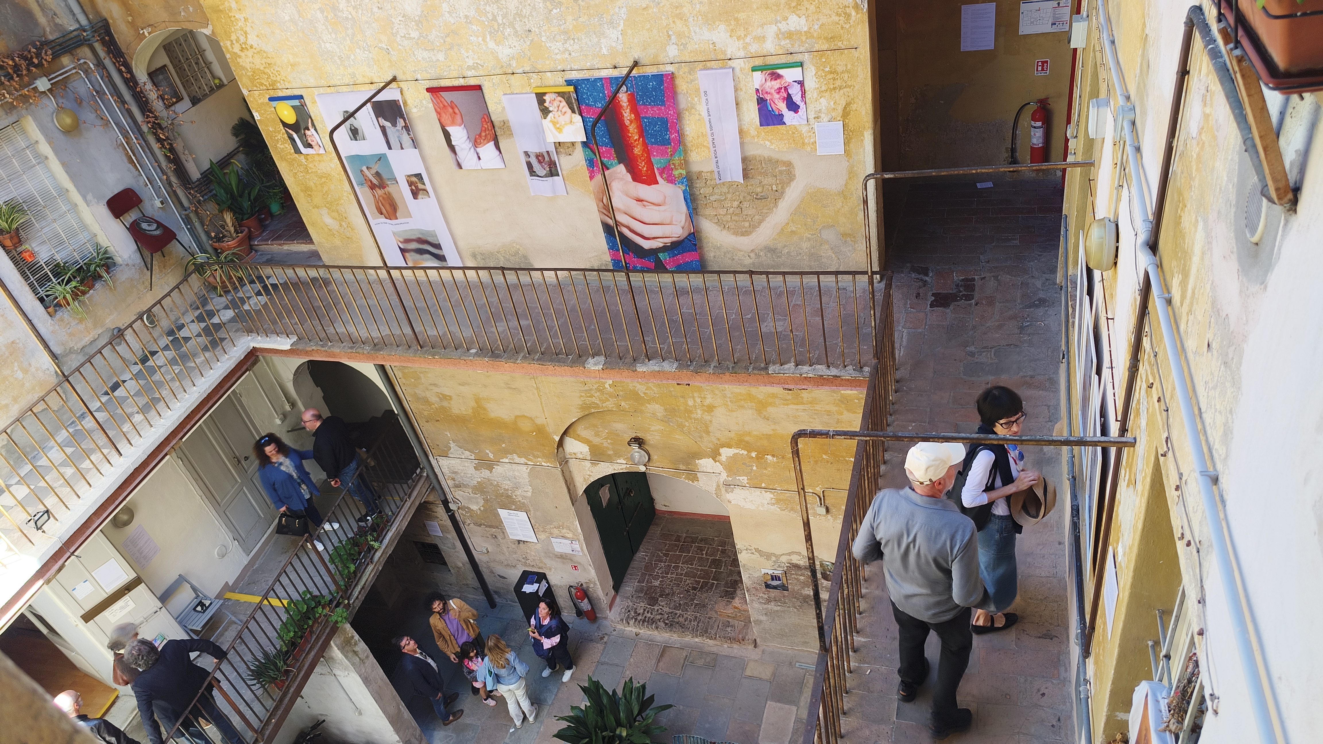 A bird's-eye view of the Atelier ViaDueGobbiTre's internal courtyard, with visitors spread across multiple levels and a line of colourful photographic works pinned along the upper balcony wall.