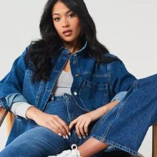 A woman sat on a chair wearing a denim jacket, grey top, and denim jeans from Very.