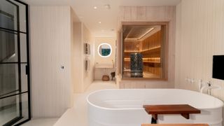luxury cream tiled bathroom with freestanding bath and bespoke built sauna in corner of room with glass walls and wooden interior