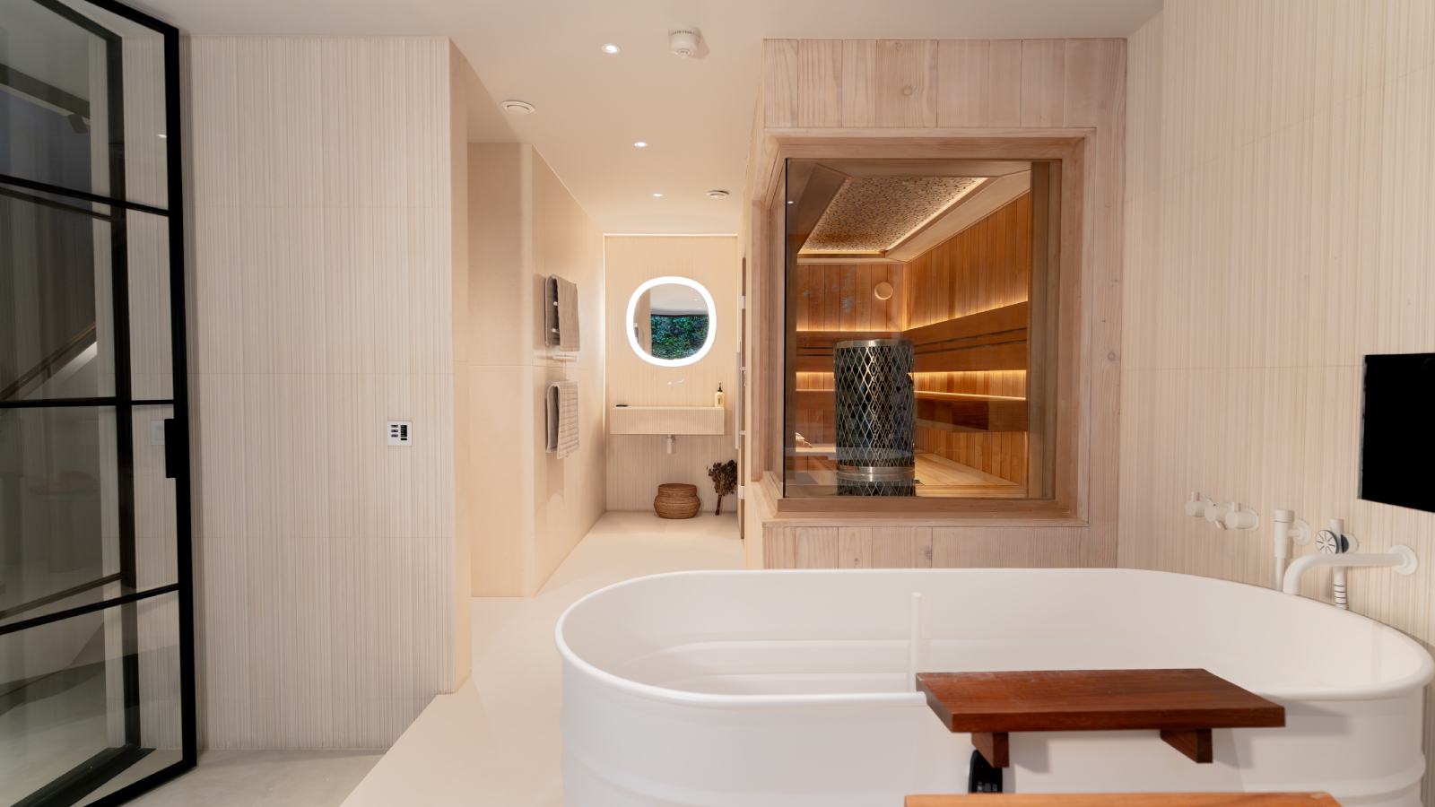 luxury cream tiled bathroom with freestanding bath and bespoke built sauna in corner of room with glass walls and wooden interior