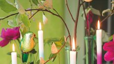 Lit taper candles against a bright green wall, pictured among branches and greenery with pink flowers and a green shiny bird decoration.