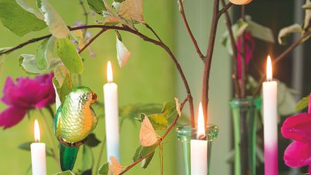 Lit taper candles against a bright green wall, pictured among branches and greenery with pink flowers and a green shiny bird decoration.