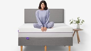 A woman with dark hair and wearing purple pajamas sits on the Purple Plus mattress