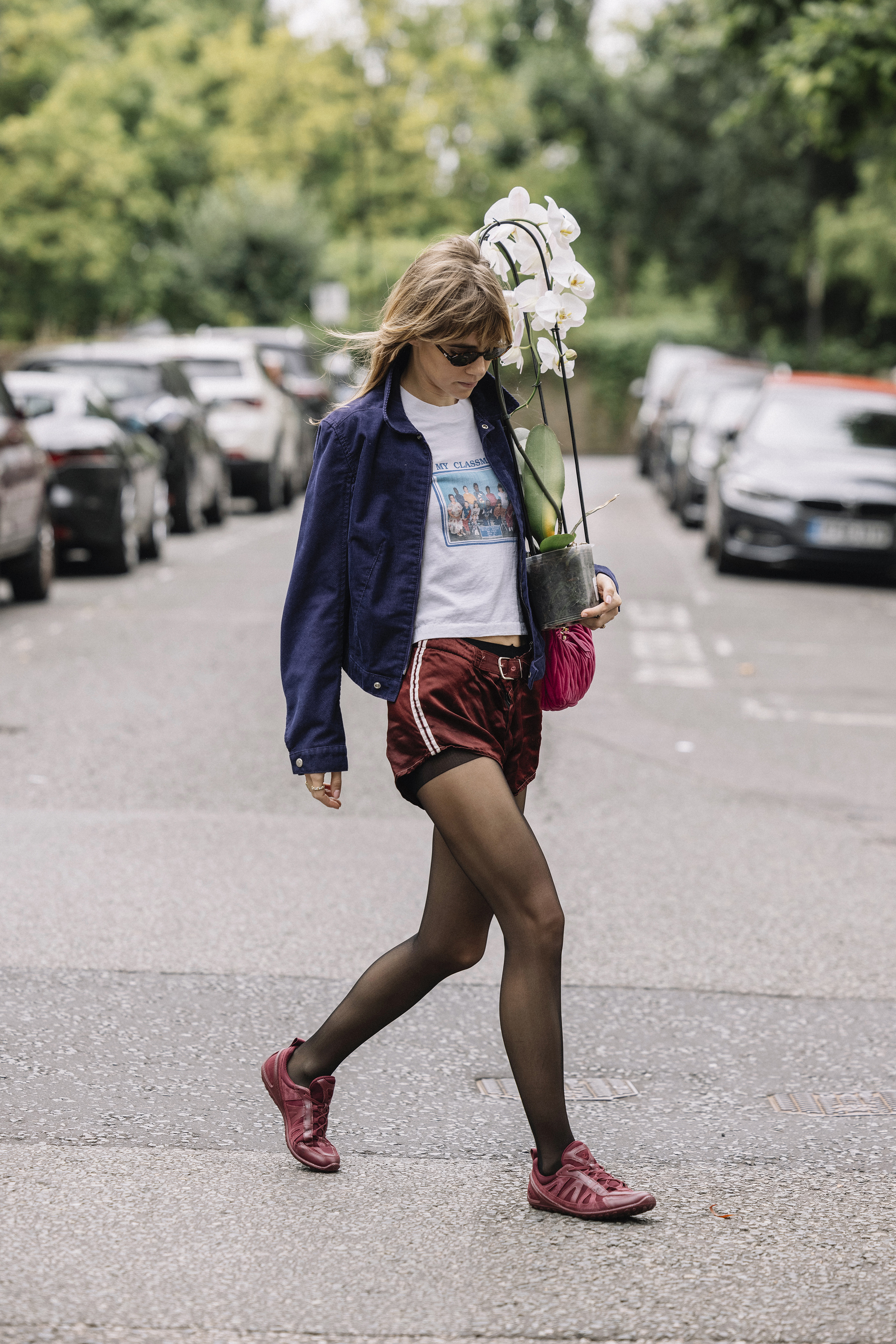 Suki Waterhouse viste una chaqueta azul marino, una camiseta blanca, pantalones cortos y zapatillas burdeos mientras lleva una planta en Londres.