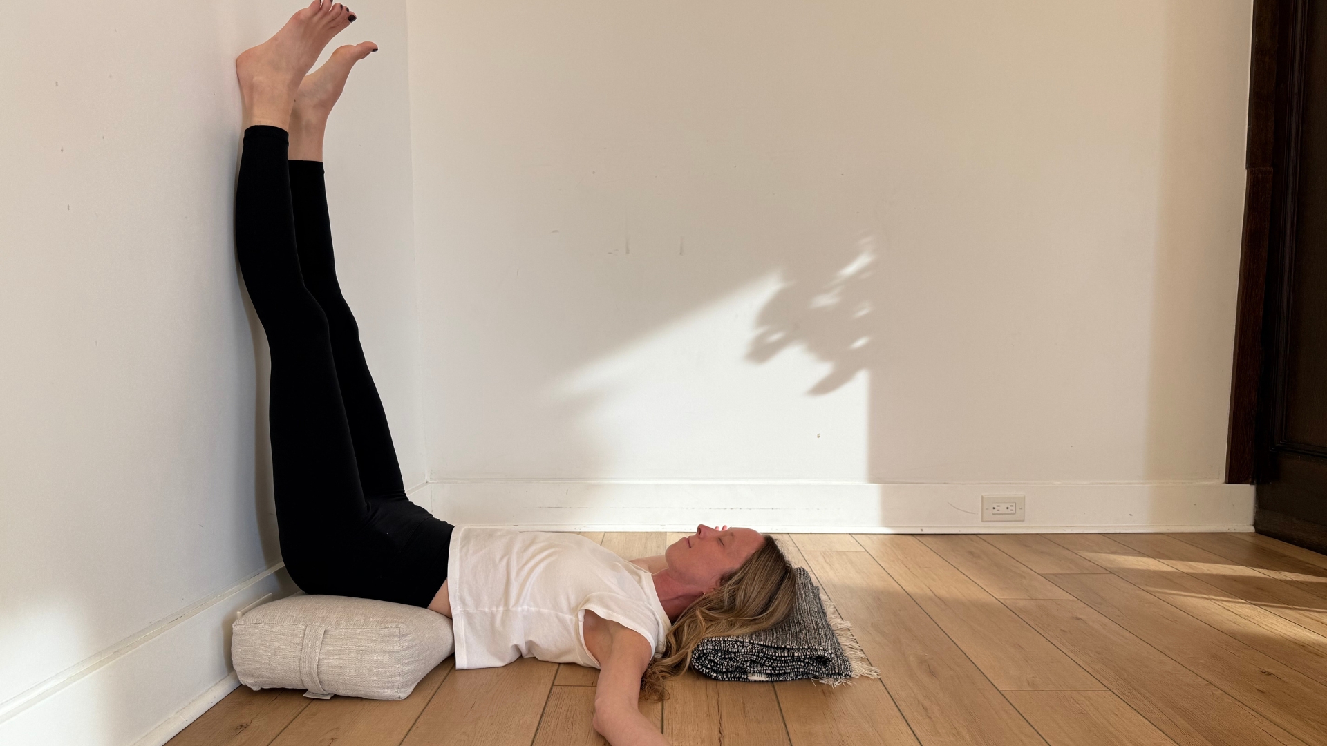 woman in legs up the wall pose on a wooden floor with white walls behind her and a cushion under her hips.
