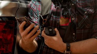 A person holding a phone and a Motorola Defy Smartphone Satellite Link attached to their backpack's strap
