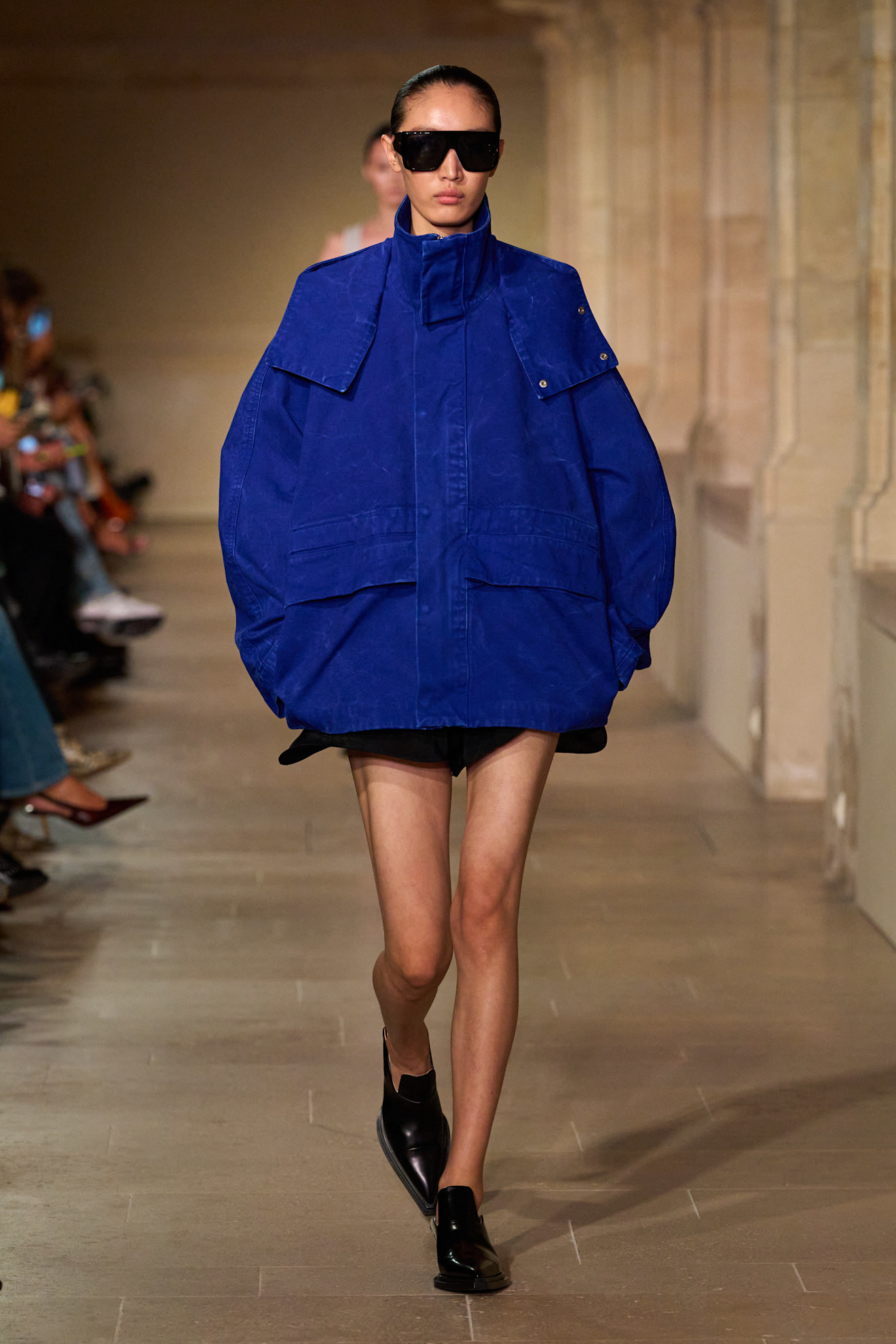 A Victoria Beckham model wearing an oversize cobalt blue funnel neck jacket layered over black shorts, black pointed toe loafers, and oversize black square sunglasses.