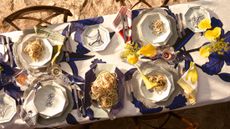 Mariana Vel&aacute;squez x Sur la Table tableware, including octagonal plates, floral table linens, and yellow flowers, pictured on a long dining table situated on a sandy beach.
