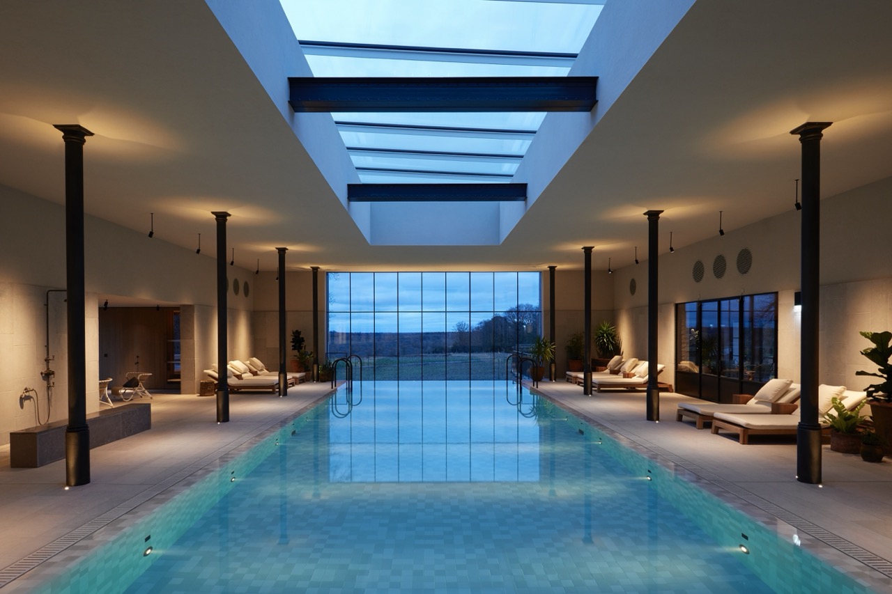 A large indoor pool beneath a skylight, lined with loungers and tall windows looking out across the fields at dusk.