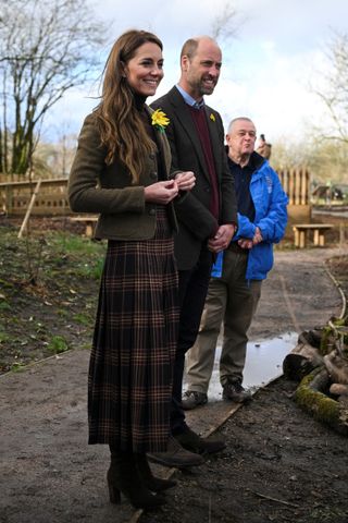 Kate Middleton wearing a long plaid skirt and short jacket with a daffodil pin smiling while standing next to Prince William on a muddy garden path