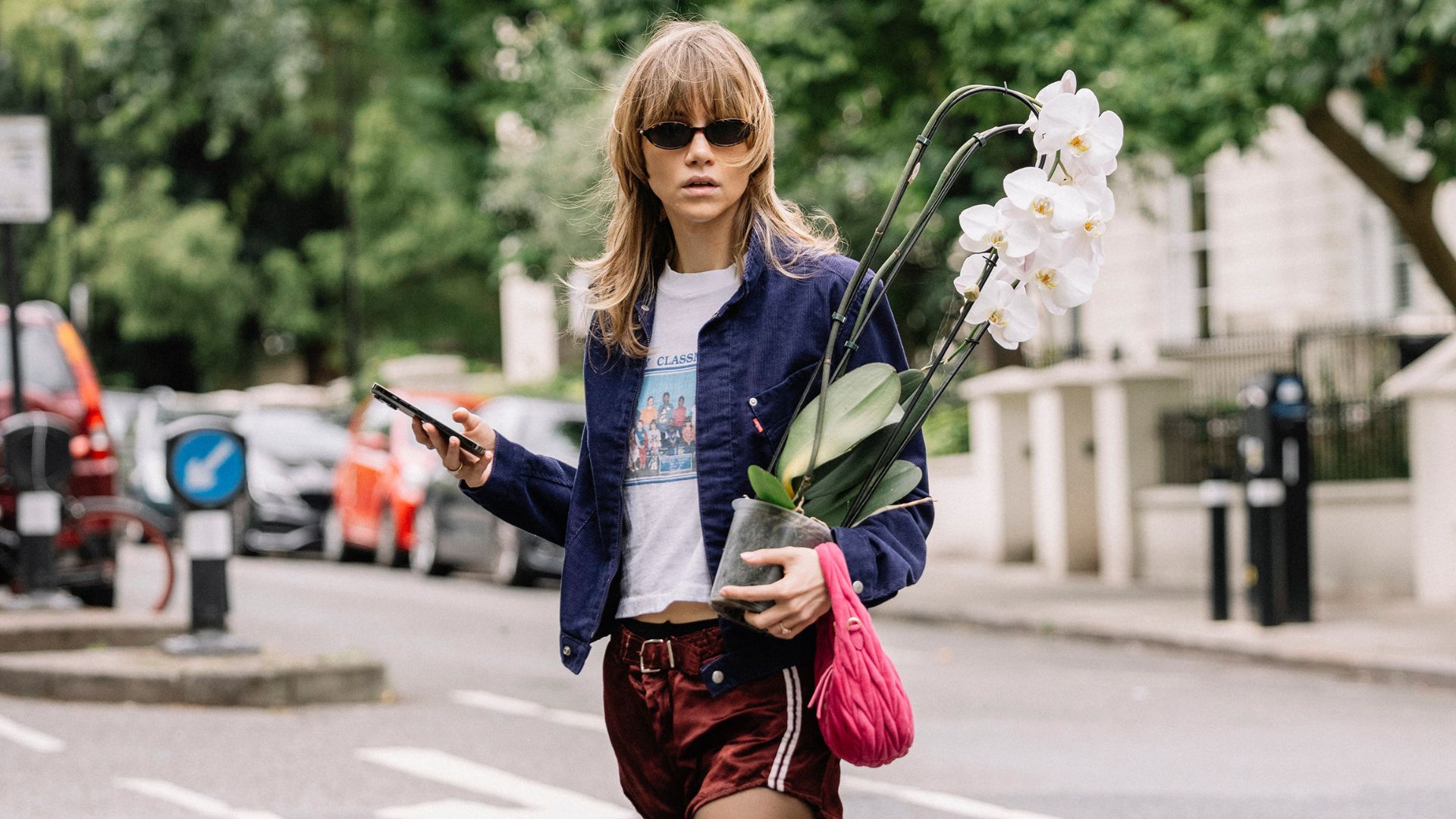 Suki Waterhouse crosses the road in London holding a large orchard over her shoulder. She wears a white graphic t-shirt, a blue bomber jacket, red satin shorts and red Ecco trainers.