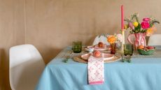 table dressed in blue tablecloth, pink and white napkins, pink plates on woven charger plates, green glassware, a vase with florals, candlesticks with pink taper candles and cutlery