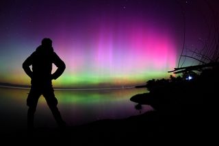A person is silhouetted by pink, purple and green northern lights on a lake shore.